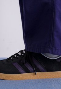 Juodas suede sportbačiai su purpurinėmis juostelėmis, reljefine guma padu ir juodais raišteliais. Dėvimi su marinų spalvos kelnėmis, matančiomis kulkšnį ir batų detales.