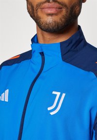 Chaqueta deportiva azul con cremallera, que presenta acentos oscuros en los hombros y un logo blanco. El tejido es suave con una textura ligeramente brillante.