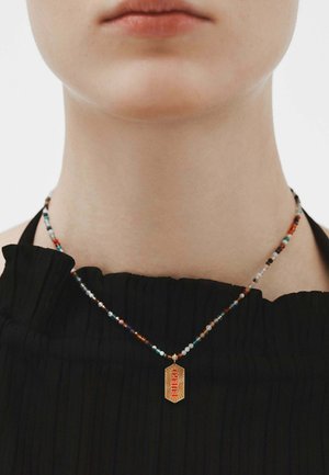 Collar de cuentas con piedras multicolores y un colgante dorado que dice "FUBO", llevado sobre una blusa negra texturizada con pliegues.