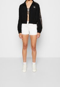 Svart bomberjacka med dragkedja, med en logotyp, långa ärmar med spetsdetaljer, kombinerad med vita shorts och sneakers.