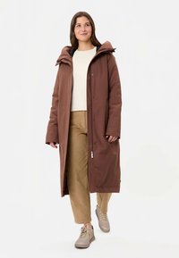 Vrouw die wandelt en een lange bruine kapmantel, beige broek, wit shirt en beige sneakers draagt tegen een effen witte achtergrond.