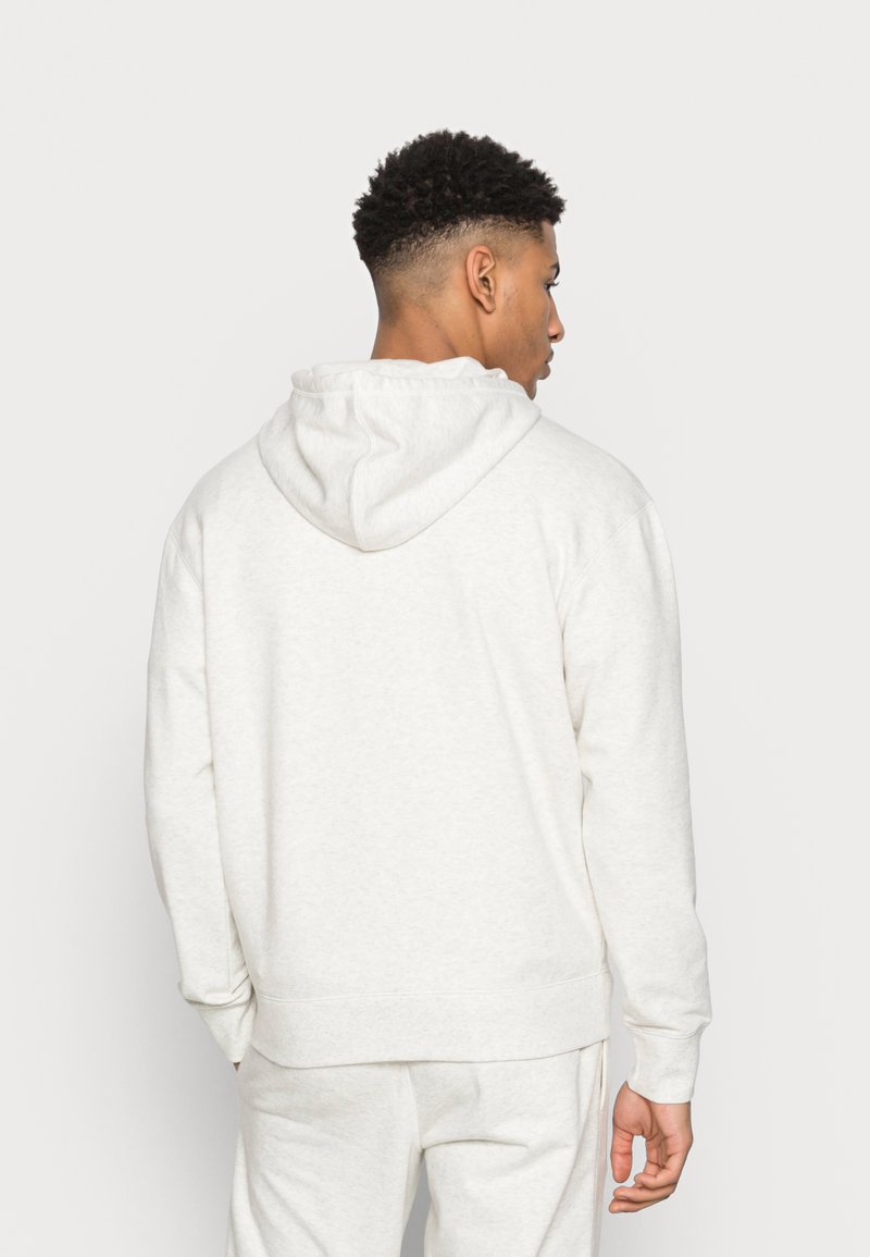Giovane uomo con capelli corti e ricci, indossa una semplice felpa con cappuccio grigia chiara e pantaloni della tuta coordinati, girato di spalle.