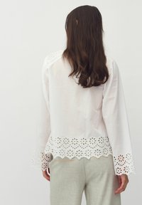 Blusa bianca a maniche lunghe con orlo scallop e ricami floreali, tessuto testurizzato e dettagli intricati sulle spalle. Abbinata a pantaloni chiari.