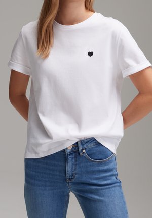 Woman wearing a plain white rolled-sleeve t-shirt with a small black heart on the left chest, paired with blue denim jeans.