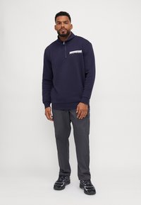 Granatowy sweatshirt z wysokim kołnierzem i pół-zamkiem, z białym logo. W połączeniu z szarymi spodniami typu cargo i czarnymi sneakersami.