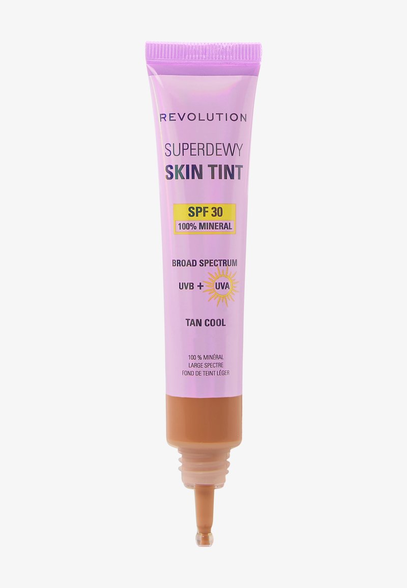 Tube violet de Revolution Superdewy Skin Tint, étiqueté « Tan Cool », avec un fond marron, affichant un SPF 30 et des détails sur la protection solaire minérale.