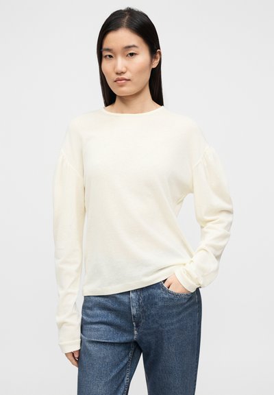 Lahka kremna zgornja majica z dolgimi rokavi in okroglim izrezom ter napihnjenimi rameni, izdelana iz mehkega blaga. Združena z modrimi jeans hlačami.