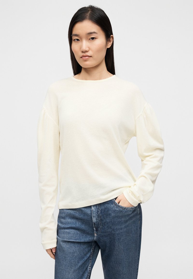 Haut crème clair à manches longues avec un col rond et des épaules bouffantes, en tissu doux. Assorti à un jean en denim bleu.