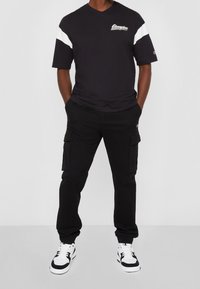 Camiseta negra con rayas blancas en los hombros y un logo, combinada con pantalones cargo negros y zapatillas en blanco y negro, todo con un ajuste relajado.
