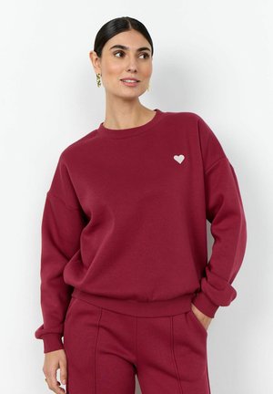 Woman wearing matching burgundy sweatshirt and pants, with small white heart detail on sweatshirt, standing against a white background.