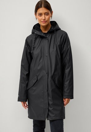 Kobieta ubrana w czarny, wodoodporny, długi płaszcz przeciwdeszczowy z kapturem, przednimi kieszeniami i zapięciem na zatrzaski, stojąca na jednolitym tle.