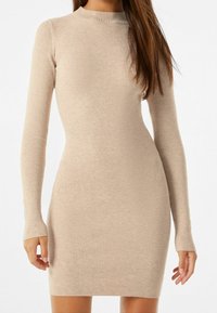 Robe en tricot côtelé de couleur beige neutre, avec des manches longues et un col haut, présentant une silhouette ajustée. Texture douce sur l'ensemble.