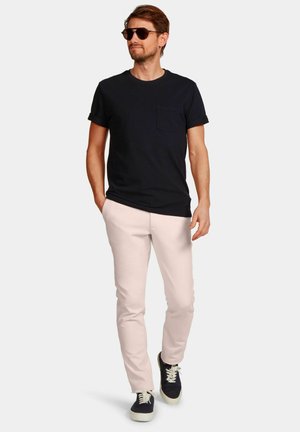 Man die een zwart T-shirt met zakken draagt, lichtroze broek, zwarte sneakers met witte zolen en een zonnebril, lopend met één hand in de zak.
