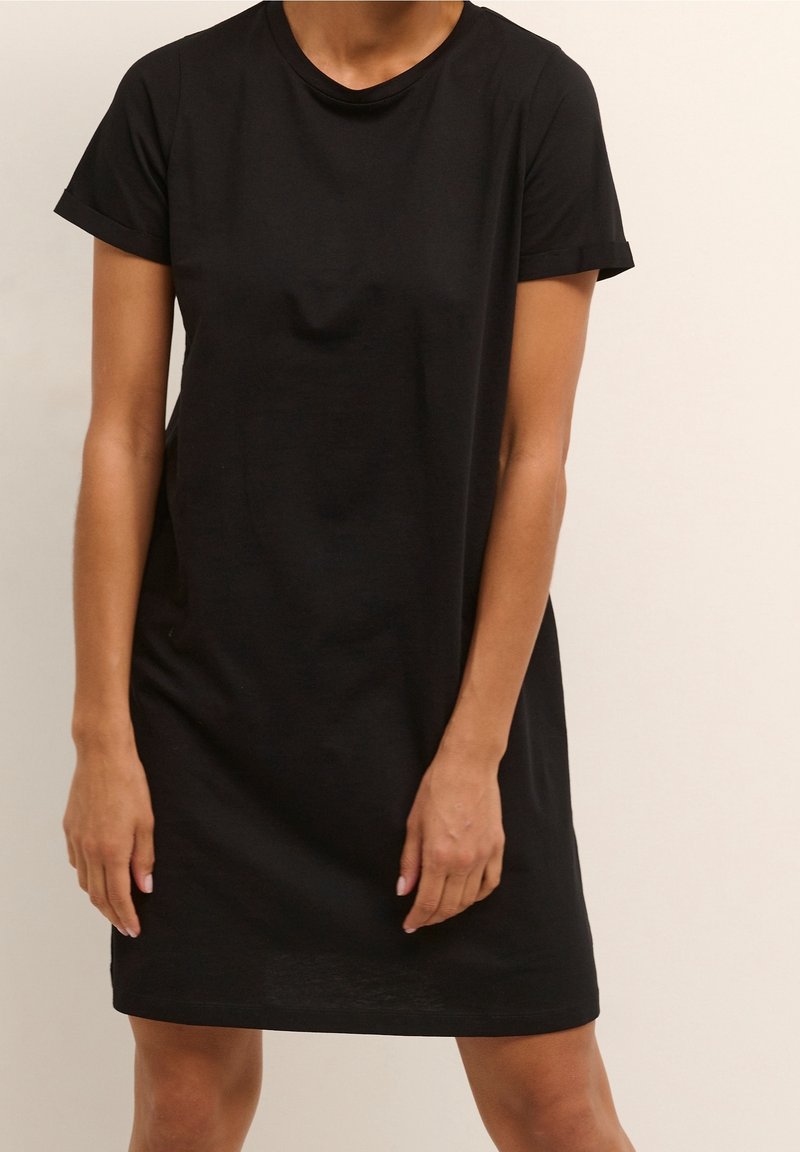 Personne portant une robe noire simple à manches courtes, debout devant un fond beige uni.