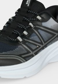 Gros plan d'une chaussure de sport noire et grise avec des lacets à motifs et une semelle blanche épaisse.