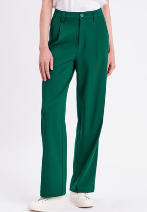 Femme portant un pantalon vert taille haute à jambes larges, associé à des baskets blanches et un haut blanc rentré, debout devant un fond blanc.
