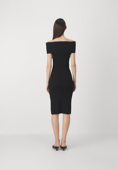 Rochie neagră din tricot cu model texturat, cu decolteu off-the-shoulder, siluetă ajustată și lungime până la genunchi. Pur cântată cu pantofi cu toc deschis la vârf, negri.