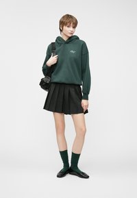 Green hooded sweatshirt with embroidered logo, paired with a pleated black skirt. Accessories include black shoulder bag and dark green knee-high socks.