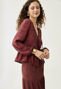 Blusa de estampado floral en color burdeos con mangas abullonadas, dobladillo con volantes y lazo en la parte delantera. Combinada con una falda texturizada a juego. Acabado con una pulsera dorada.