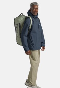 Giacca impermeabile blu marina, pantaloni cargo beige e scarpe da trekking grigie. Grande zaino verde con dettagli neri e logo sul lato.