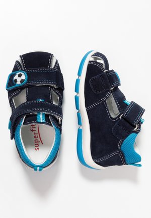Marineblå ruskindssko med mesh-detaljer, justerbare velcroremme, fodbolddetalje og kontrasterende lyseblå såler og syninger.