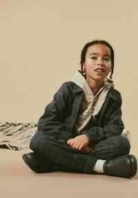 Barn som har på sig en grå rutmönstrad jacka över en beige hoodie och mörka byxor, med svarta träskor och vita strumpor, sitter mot en neutral bakgrund.