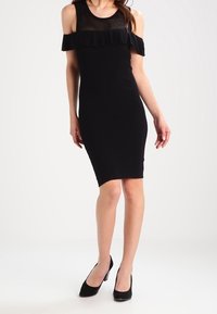 Vestito nero aderente con design off-shoulder, pannello in rete e dettagli con volant sul collo. Abbinato a scarpe con tacchi alti nere.