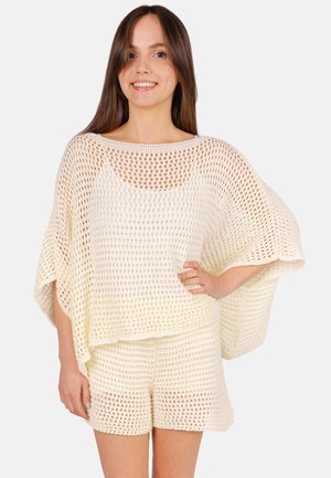 Young woman standing smiling wearing cream-colored loose knit top and matching shorts against plain background.
