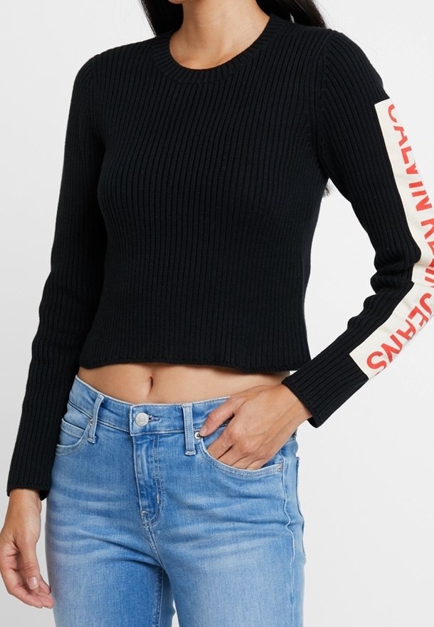 Femme portant un crop top à manches longues côtelé noir avec le texte "Calvin Klein Jeans" en rouge sur des bandes blanches le long des manches, assorti à un jean bleu clair.