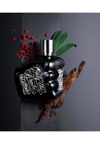 Bouteille en verre noir en forme de poing, bouchon argent, inscription blanche "Only the Brave Tattoo", accompagnée de baies rouges et de feuilles vertes.