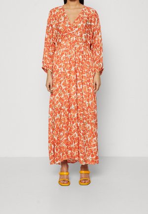 Femme portant une longue robe fluide à motifs floraux orange et blancs, avec des sandales à talons ouvertes jaunes, debout devant un fond uni.