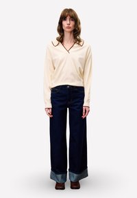 Vrouw met schouderlang bruin haar, gekleed in een crèmekleurige trui met een bruine kraag, brede donkerblauwe jeans met omgeslagen zomen en bruine schoenen.