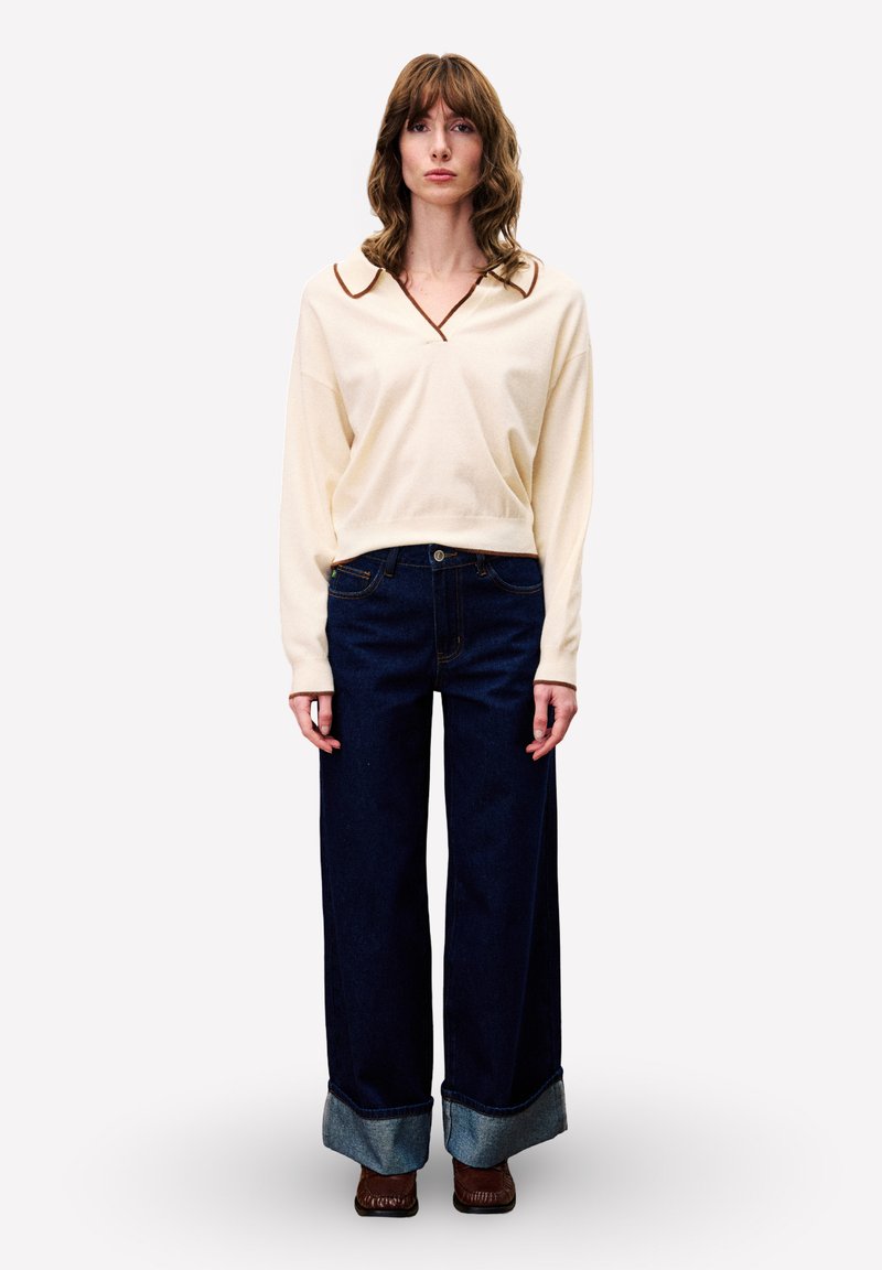 Vrouw met schouderlang bruin haar, gekleed in een crèmekleurige trui met een bruine kraag, brede donkerblauwe jeans met omgeslagen zomen en bruine schoenen.