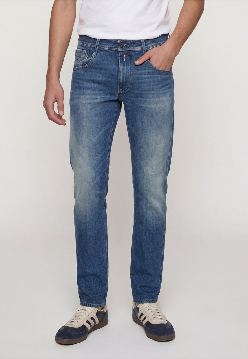 Homme portant un jean bleu délavé, un t-shirt blanc et des baskets bleues et blanches, une main dans la poche, debout devant un fond uni.