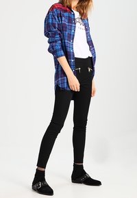 Camicia a maniche lunghe a quadri blu e rossi, indossata sopra una t-shirt bianca con grafica. Pantaloni neri slim con tasche con zip e stivaletti.