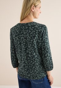 T-shirt à manches longues, de couleur vert foncé, avec un motif léopard. Texture douce, encolure ronde et coupe décontractée avec des manches légèrement bouffantes.