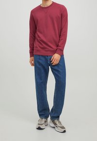 Felpa bordeaux in morbido tessuto con colletto a girocollo e polsini a costine, abbinata a jeans in denim blu larghi e sneakers grigie.