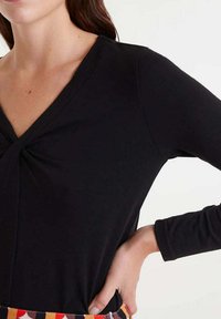 Top negro de manga larga con escote en V y un detalle retorcido en la parte delantera. Textura suave, diseño ajustado y tela lisa.