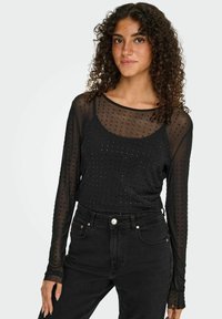 Sort langærmet top med gennemsigtigt stof og prikkede dekorationer. Har en tætsiddende pasform og rund halsudskæring, kombineret med højtaljede sorte jeans.
