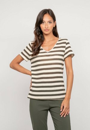 Femme aux cheveux longs et foncés portant un haut rayé olive et blanc à manches courtes et un pantalon olive, debout avec une main sur la hanche.