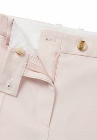 Jasnoróżowe spersonalizowane spodnie zawierają zapięcie na guzik, zamek błyskawiczny oraz teksturowany materiał. Wnętrze podszewki jest białe, co uwydatnia staranne detale szycia.