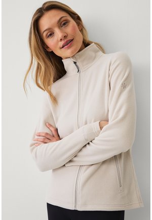 Vrouw met blond haar, gekleed in een lichtbeige fleecejack met rits, staand met gekruiste armen tegen een effen witte achtergrond.