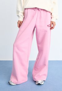 Pantalons de survêtement larges roses avec une texture douce, une taille élastique et un détail à cordon de serrage. Présente un petit logo brodé sur le côté.