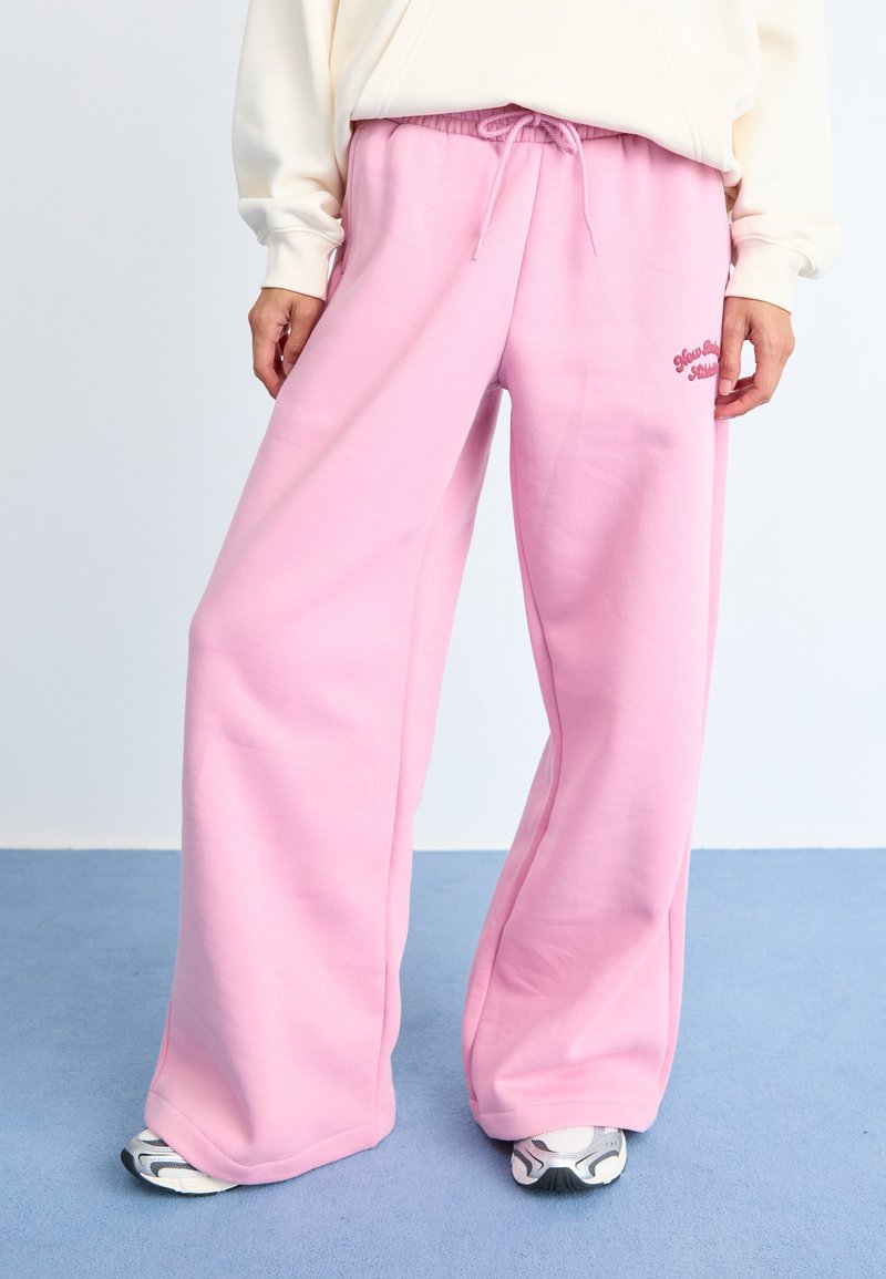 Pantalons de survêtement larges roses avec une texture douce, une taille élastique et un détail à cordon de serrage. Présente un petit logo brodé sur le côté.