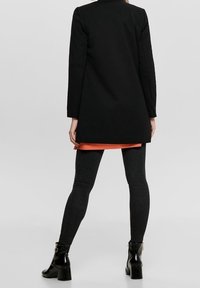 Svart lång kappa med hög krage, bärs över en ljus orange topp, kombinerad med svarta skinny jeans och ankelboots. Mjukt tyg, skräddarsydd passform.