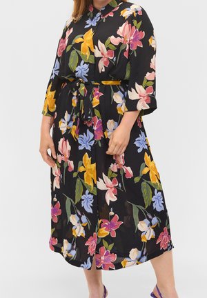 Nainen pukeutuneena mustaan midi-mittaisen mekkoon, jossa on värikäs kukkakuvio, napitettu edestä, kolmen neljänneksen hihat ja vyötäröllä solmittu samaa kangasta oleva vyö.