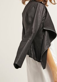 Black leather jacket with a cropped design, featuring a silver zipper and an asymmetrical hem. Smooth texture and minimalistic style.