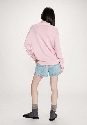 Femme aux longs cheveux foncés vue de dos, portant un pull rose ample, un short en jean clair, des chaussettes grises et des chaussures noires sans lacets.