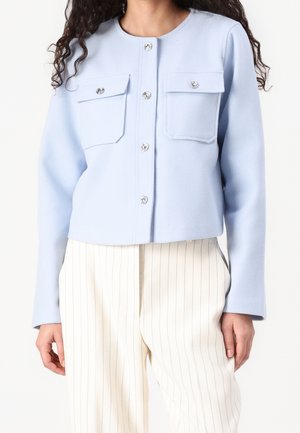 Femme portant une veste boutonnée bleu clair avec deux poches poitrine et un pantalon large crème à fines rayures verticales.