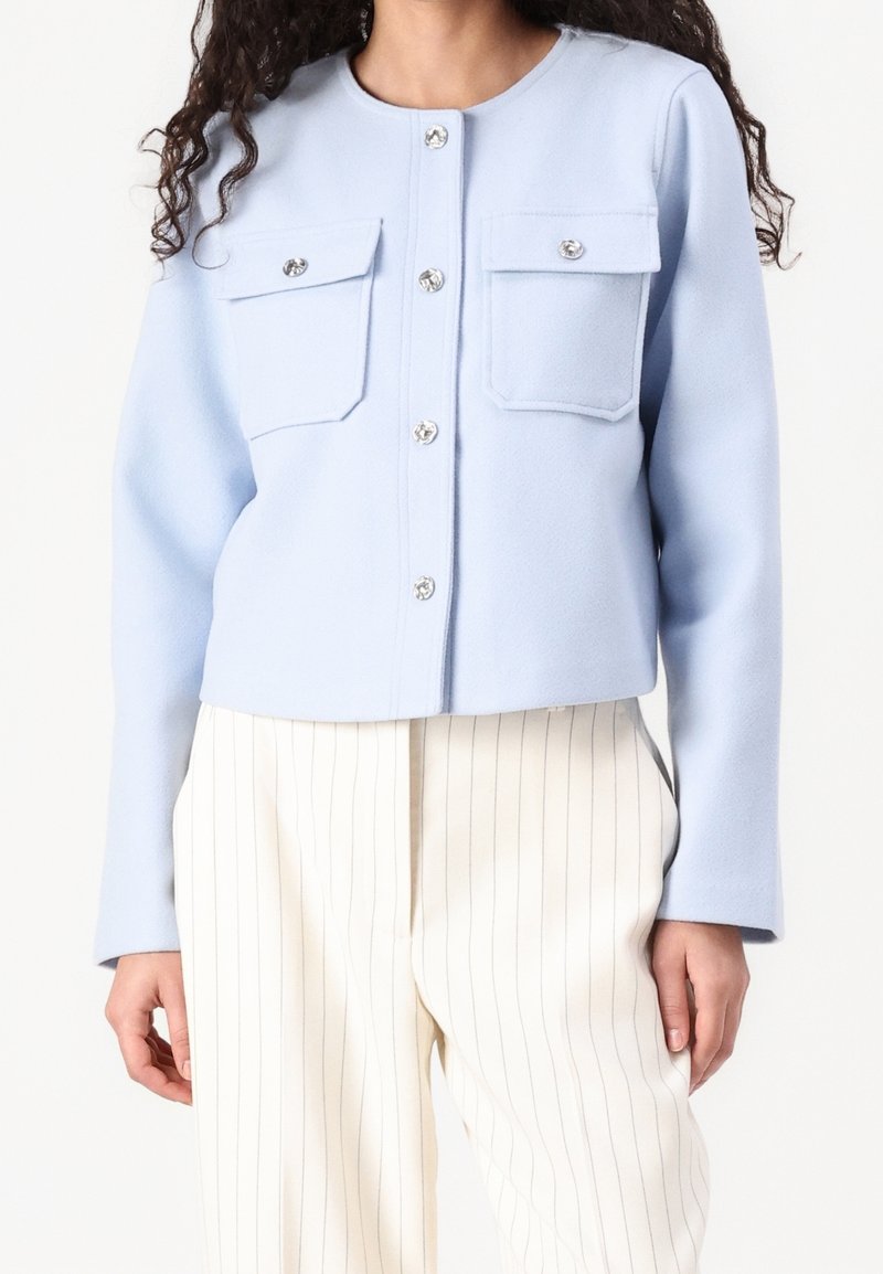 Femme portant une veste boutonnée bleu clair avec deux poches poitrine et un pantalon large crème à fines rayures verticales.